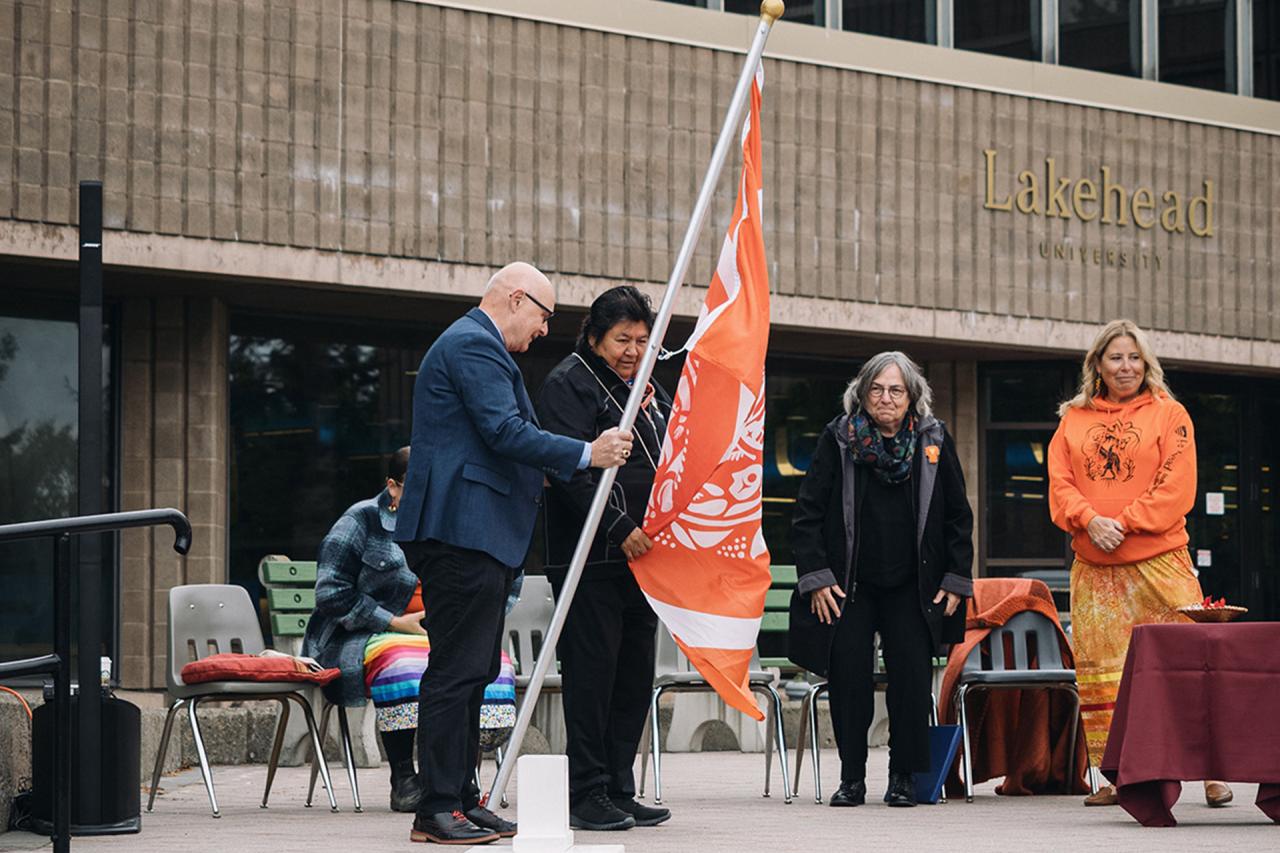 Lorne Clifford, Director of Security Services at Lakehead, and Elder Catherine McGuire raise the Survivors' Flag outside of Lakehead's Agora.