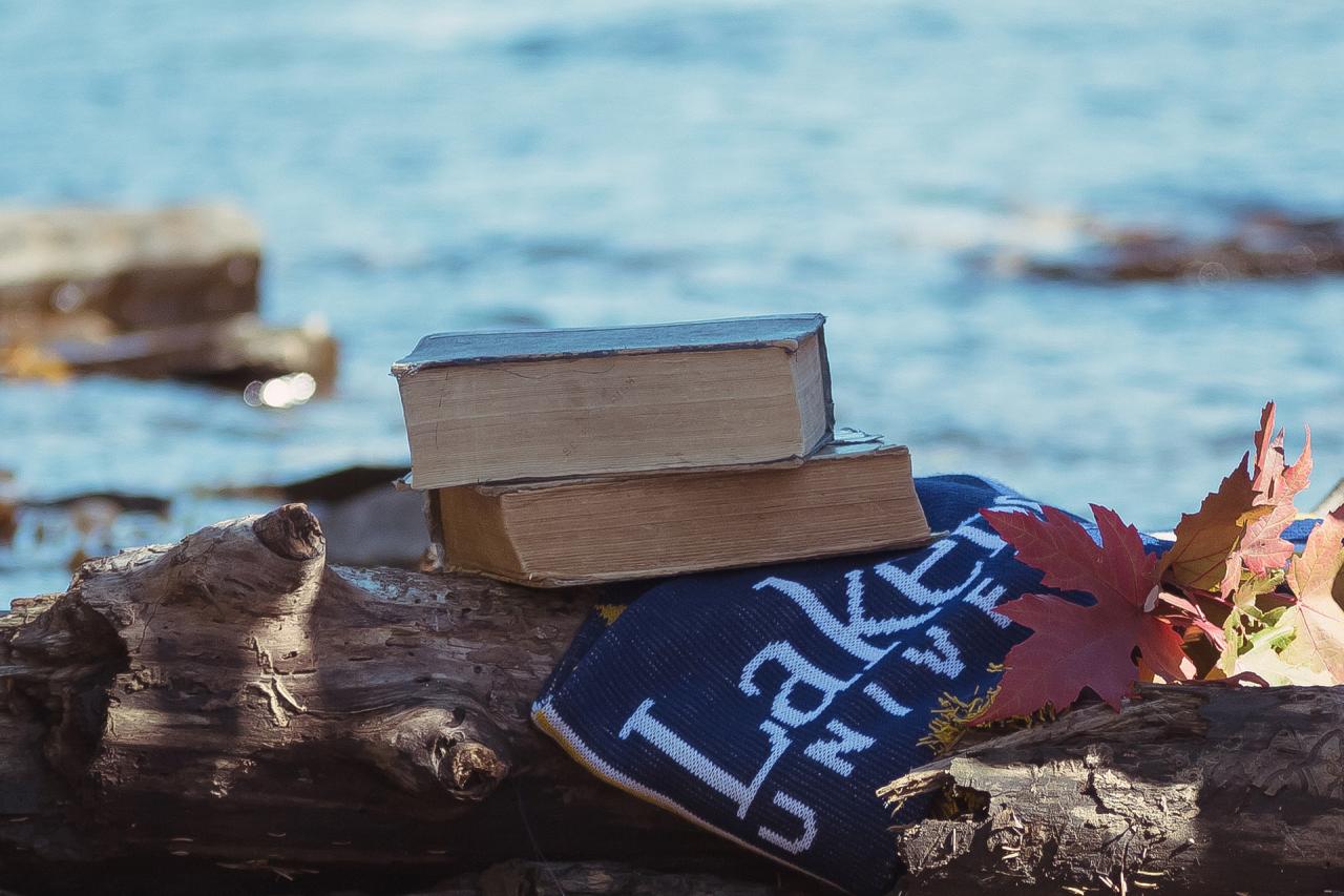 Image of Lakehead University Scarf, Maple Leaves, and books on top of a rock with water in the background 